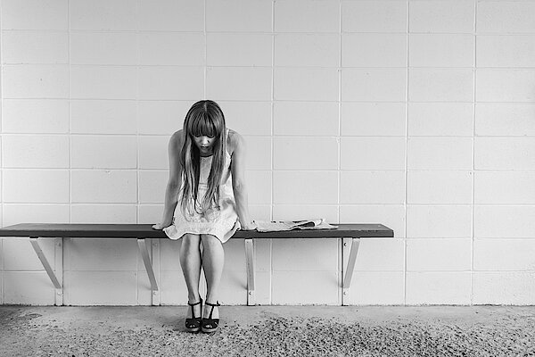 Schwarzweiß Aufnahme einer jungen Frau. Sie sitzt traurig und alleine auf einer Bank.